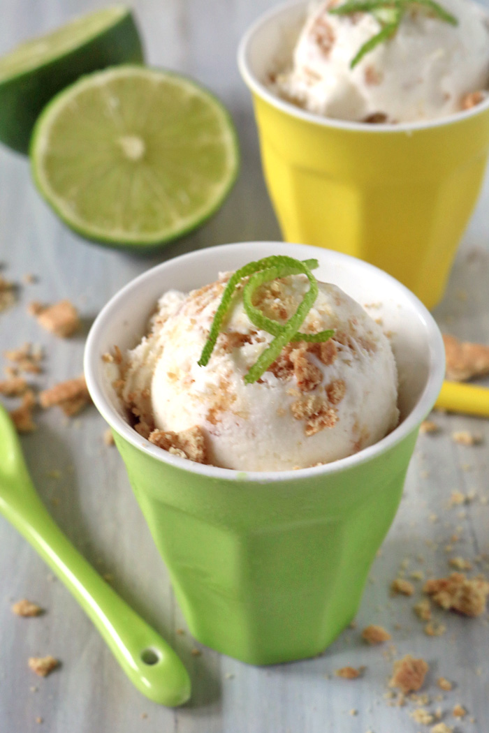 A scoop of ice cream sprinkled with crumbs and topped with lime zest sits in a green cup, with a green spoon nearby. A yellow cup with more ice cream and a halved lime are in the background.