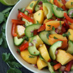 A bowl of fresh mango salsa with chopped mango, cucumber, red pepper, red onion, and cilantro. Lime and cilantro leaves are beside the bowl on a dark surface.