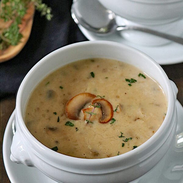 A white bowl filled with creamy mushroom soup, garnished with mushroom slices and chopped herbs, sits on a matching saucer with a spoon beside it. Another bowl of soup and fresh herbs are visible in the background.