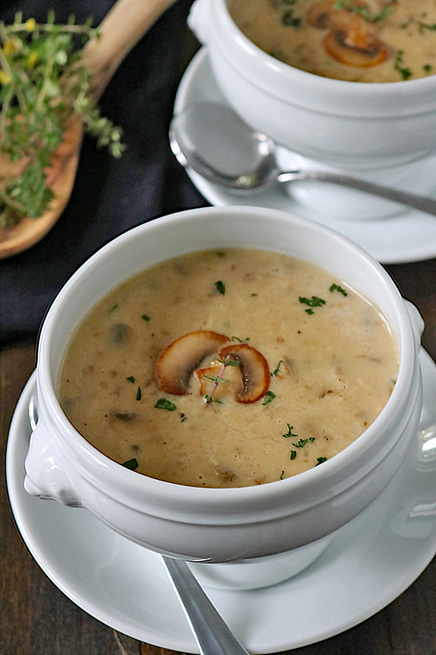 A white bowl filled with creamy mushroom soup, garnished with mushroom slices and chopped herbs, sits on a matching saucer with a spoon beside it. Another bowl of soup and fresh herbs are visible in the background.