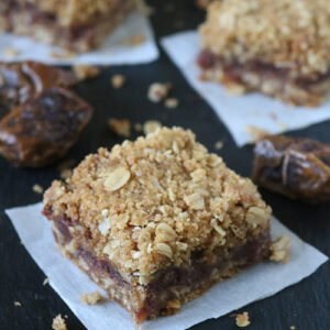 Three square oat date bars with crumbly topping sit on parchment pieces, surrounded by scattered oats and date pieces on a dark surface.