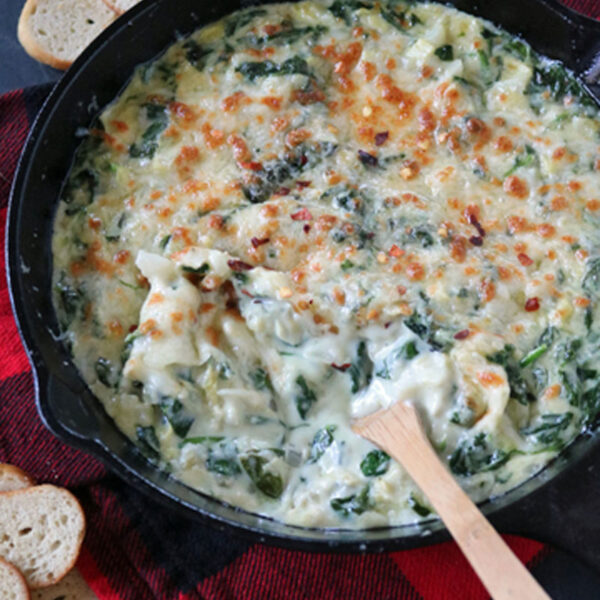 A cast iron skillet filled with creamy, baked spinach and cheese dip sits on a red plaid cloth, surrounded by toasted bread slices. A wooden spoon rests in the dip, which has a golden, bubbly top.