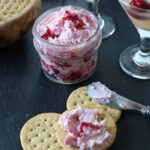 A jar of cranberry cheese spread with two cocktails, some round crackers, and a knife spreading the mixture on a cracker, all on a dark surface.