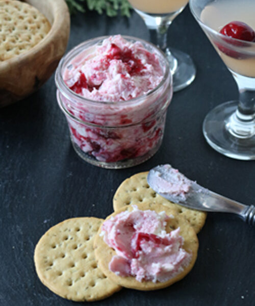 A jar of cranberry cheese spread with two cocktails, some round crackers, and a knife spreading the mixture on a cracker, all on a dark surface.