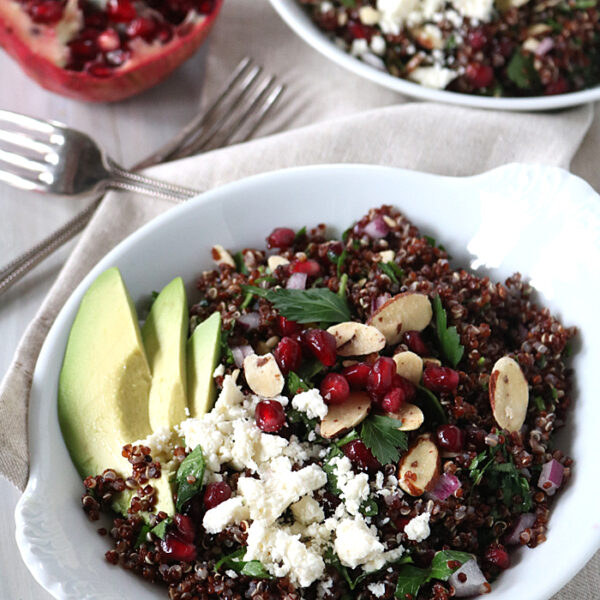 A white bowl filled with quinoa salad topped with feta cheese, pomegranate seeds, sliced almonds, herbs, and avocado slices. Another bowl and a halved pomegranate are in the background on a light table.