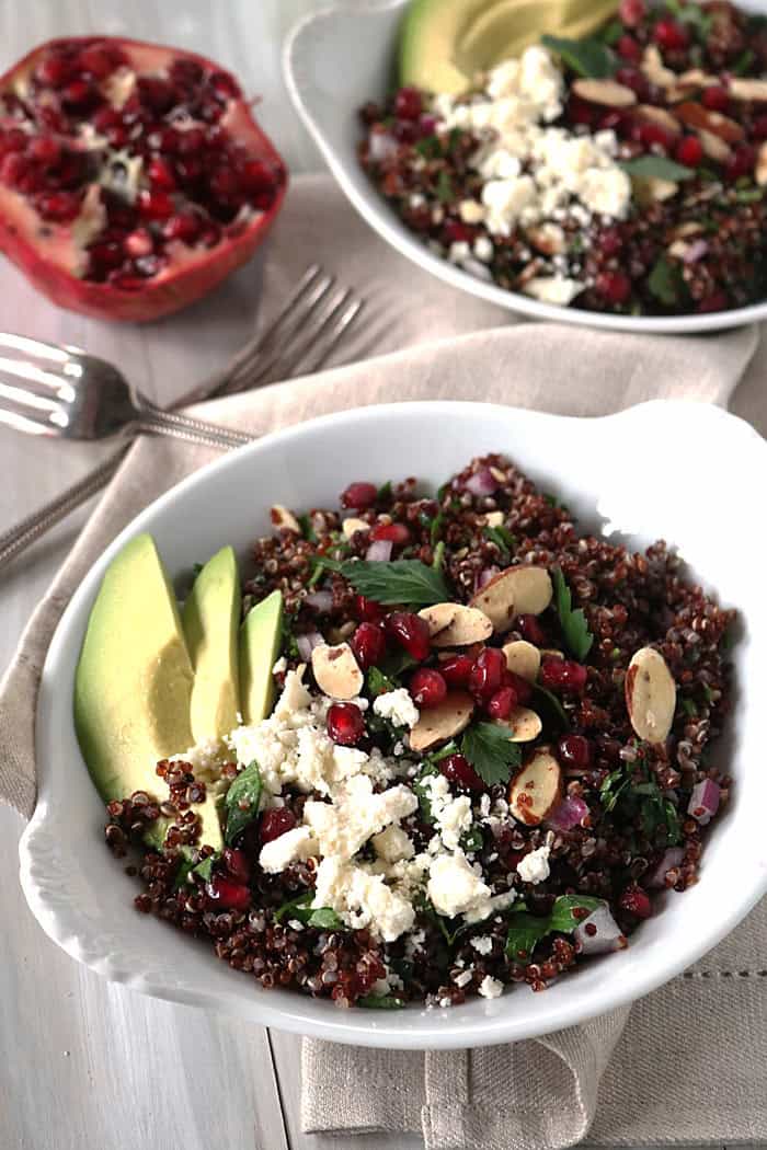 A bowl of holiday quinoa crunch salad topped with crumbled feta, pomegranate seeds, almond slices, and parsley, with sliced avocado on the side. Another similar bowl and a halved pomegranate are in the background.