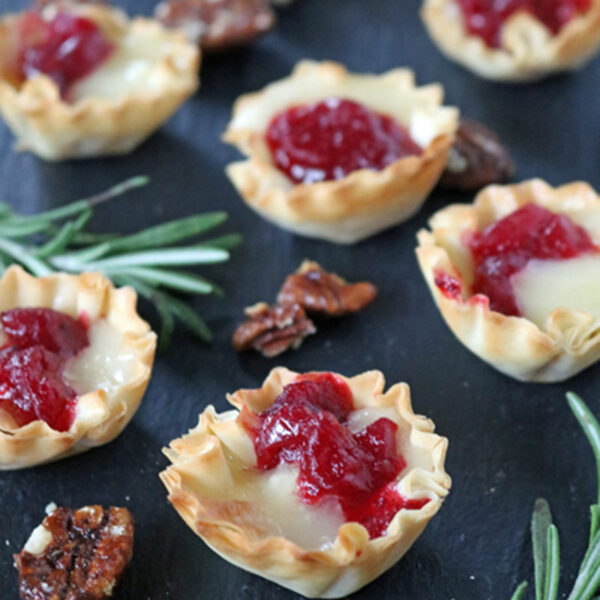 Mini phyllo pastry cups filled with melted cheese and red cranberry sauce are arranged on a dark slate surface, garnished with sprigs of fresh rosemary and pieces of candied pecans.