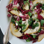 A white plate with a salad of sliced pears, radicchio, parsley, crumbled feta cheese, and chopped pistachios, served with a silver spoon on the side.