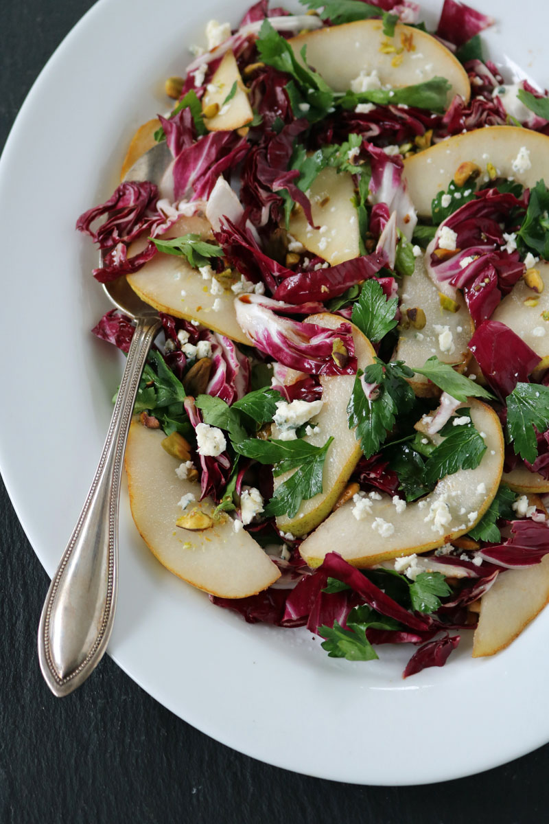 A white plate with a salad of sliced pears, radicchio, parsley, crumbled feta cheese, and chopped pistachios, served with a silver spoon on the side.