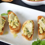 Four pieces of toasted bread topped with a chunky greenish-white spread and garnished with shaved cheese, served on a white plate with fresh basil and a bowl of spread in the background.