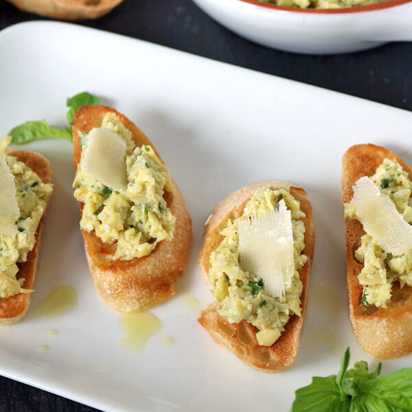 Four pieces of toasted bread topped with a chunky greenish-white spread and garnished with shaved cheese, served on a white plate with fresh basil and a bowl of spread in the background.