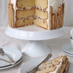A layered vanilla cake with chocolate chips and white frosting is decorated with rolled wafer cookies around the sides. A slice is served on a plate beside the cake, which sits on a white cake stand.