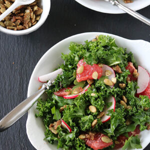 A bowl of kale salad with sliced radishes, grapefruit segments, and crunchy seeds. A spoon rests in the bowl. Another smaller white dish with seeds and a spoon sits nearby on a dark surface.