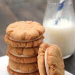 A stack of peanut butter sandwich cookies sits on a napkin next to a glass bottle of milk with a blue-striped straw. The cookies have a crisscross pattern on top.