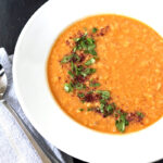 A bowl of orange lentil soup garnished with chopped cilantro and spices, served on a white plate with a spoon and napkin beside it. Small dishes of herbs and spices are partially visible in the background.