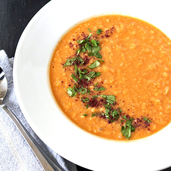 A bowl of orange lentil soup garnished with chopped cilantro and spices, served on a white plate with a spoon and napkin beside it. Small dishes of herbs and spices are partially visible in the background.
