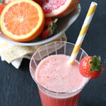 A glass of pink fruit juice with a yellow-and-white striped straw and a strawberry garnish. Behind it, a plate holds a halved grapefruit, an orange slice, and strawberries.