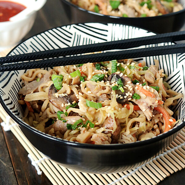 A bowl of stir-fried noodles with vegetables, mushrooms, and sesame seeds, garnished with chopped green onions. Black chopsticks rest on the bowl, and a small dish of red sauce is in the background.