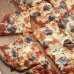 Chicago-style thin crust sausage pizza, cut into square slices and topped with melted cheese and savory sausage, sits on a wooden board. One slice is pulled away from the rest.