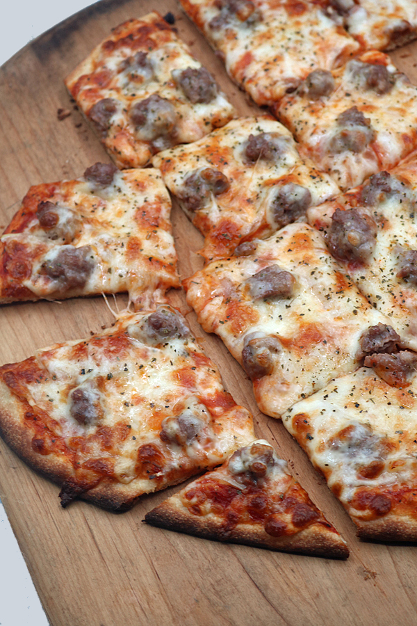 Chicago-style thin crust sausage pizza, cut into square slices and topped with melted cheese and savory sausage, sits on a wooden board. One slice is pulled away from the rest.