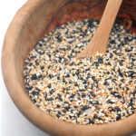 A wooden bowl filled with a mixture of black and white sesame seeds and a wooden spoon resting inside.