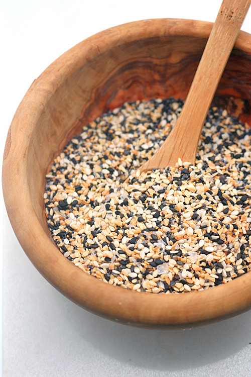 A wooden bowl filled with a mixture of black and white sesame seeds, with a wooden spoon resting inside the bowl.