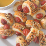 A plate of pigs in a blanket topped with everything bagel seasoning, served with a bowl of yellow mustard dipping sauce.
