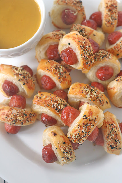 A plate of puff pastry-wrapped mini sausages sprinkled with seasoning, served with a bowl of yellow mustard dipping sauce.