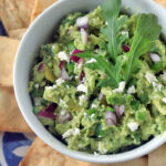 A bowl of guacamole garnished with arugula, red onion, and crumbled cheese, surrounded by pita chips on a blue and white patterned plate.