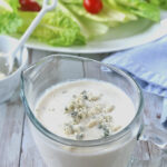 A glass measuring cup filled with creamy blue cheese dressing sits on a wooden table, with crumbled blue cheese on top. In the background, a plate of fresh romaine lettuce and cherry tomatoes is visible.