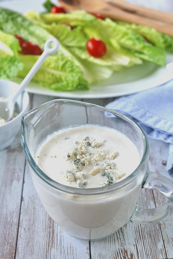 A glass pitcher filled with creamy blue cheese dressing, topped with blue cheese crumbles, sits on a rustic white table. In the background, fresh romaine lettuce and cherry tomatoes are arranged on a plate.
