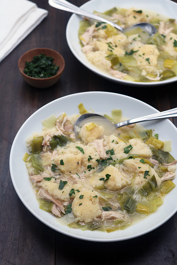 Two white bowls filled with chicken and dumplings soup, featuring chunks of chicken, dumplings, celery, and leeks, topped with chopped herbs. Spoons rest in each bowl, with a small bowl of herbs nearby.