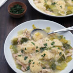 Two white bowls filled with chicken and dumplings soup, featuring chunks of chicken, dumplings, celery, and broth, garnished with chopped herbs. Spoons rest in the bowls, with a small bowl of herbs beside them.