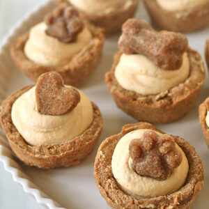Mini tart-like treats with light tan filling, topped with dog-shaped biscuits, including hearts, bones, and paw prints, arranged on a white plate.