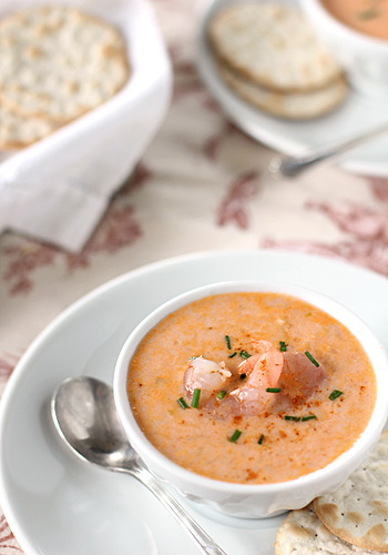 A bowl of creamy shrimp bisque garnished with chives is served on a white plate with a spoon and crackers; more crackers are in the background on a patterned tablecloth.