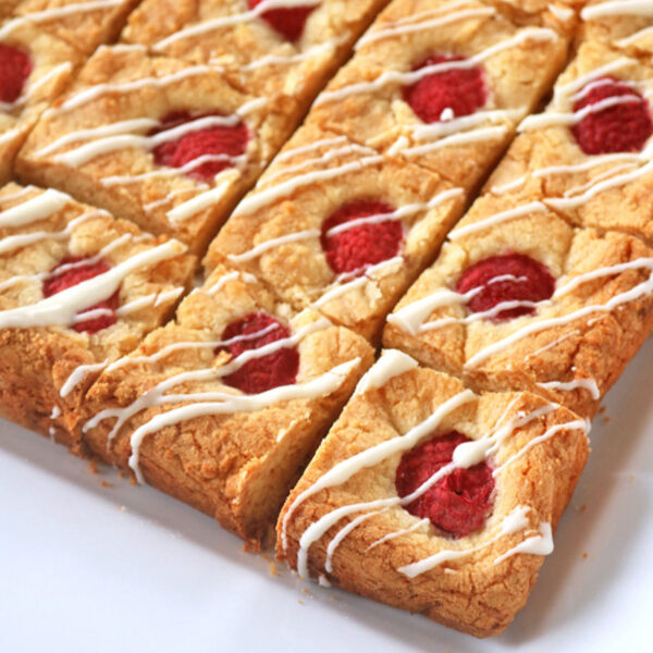 Golden blondie bars topped with fresh raspberries and drizzled with white icing, neatly cut into squares and arranged on a white plate.