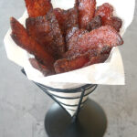 A metal cone holds crispy strips of candied bacon arranged upright in white parchment paper, set on a gray surface.