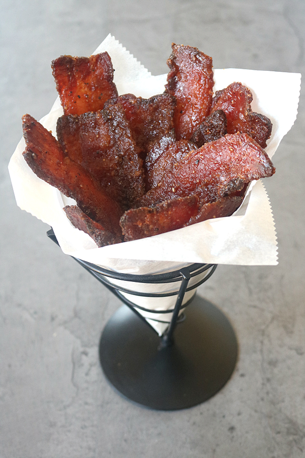 A metal cone holds crispy strips of candied bacon arranged upright in white parchment paper, set on a gray surface.