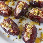 A close-up of dates stuffed with creamy cheese and chopped pistachios, arranged on a white plate and drizzled with honey.