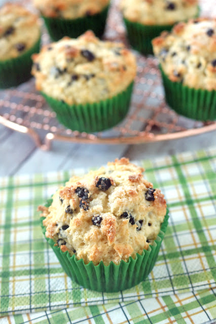 A close-up of a golden-brown muffin with dark chocolate chips in a green paper liner, sitting on a plaid cloth, with more muffins on a wire rack in the background.