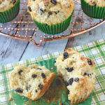 A split currant muffin with a green paper liner rests on a green plaid napkin, with whole muffins in green liners on a cooling rack in the background. The muffins are lightly golden and studded with currants.