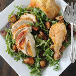 A square white plate with roasted chicken pieces, sliced and whole, served on a bed of arugula and toasted bread cubes. A serving fork and spoon rest on the side.