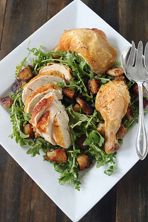 A square white plate with roasted chicken pieces, sliced and whole, served on a bed of arugula and toasted bread cubes. A serving fork and spoon rest on the side.