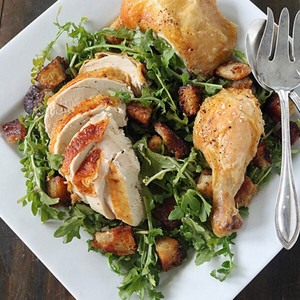 A square white plate with roasted chicken pieces, sliced and whole, on a bed of arugula and toasted bread cubes, served with metal salad utensils on a dark wooden table.