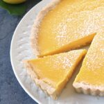 A lemon tart with a slice being taken out, topped with powdered sugar, sits on a white plate. Two fresh lemons, one cut in half, are in the background.