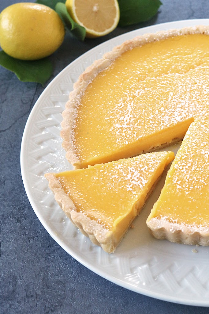 A lemon tart with a slice being taken out, topped with powdered sugar, sits on a white plate. Two fresh lemons, one cut in half, are in the background.