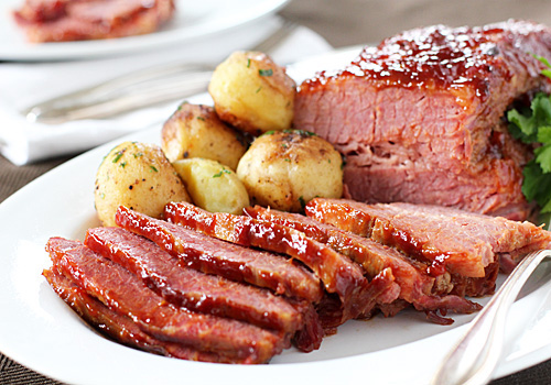 Sliced glazed corned beef served with roasted baby potatoes and garnished with herbs on a white plate. A fork and knife rest beside the plate, and another plate is visible in the background.