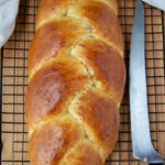 A golden-brown braided loaf of bread rests on a cooling rack next to a serrated bread knife. A white cloth is partially visible at the top of the image.