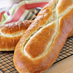 Two golden brown loaves of bread with crisp crusts rest on a black wire cooling rack, with a striped cloth in the background.