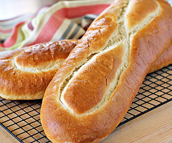 Two golden brown loaves of bread with crisp crusts rest on a black wire cooling rack, with a striped cloth in the background.